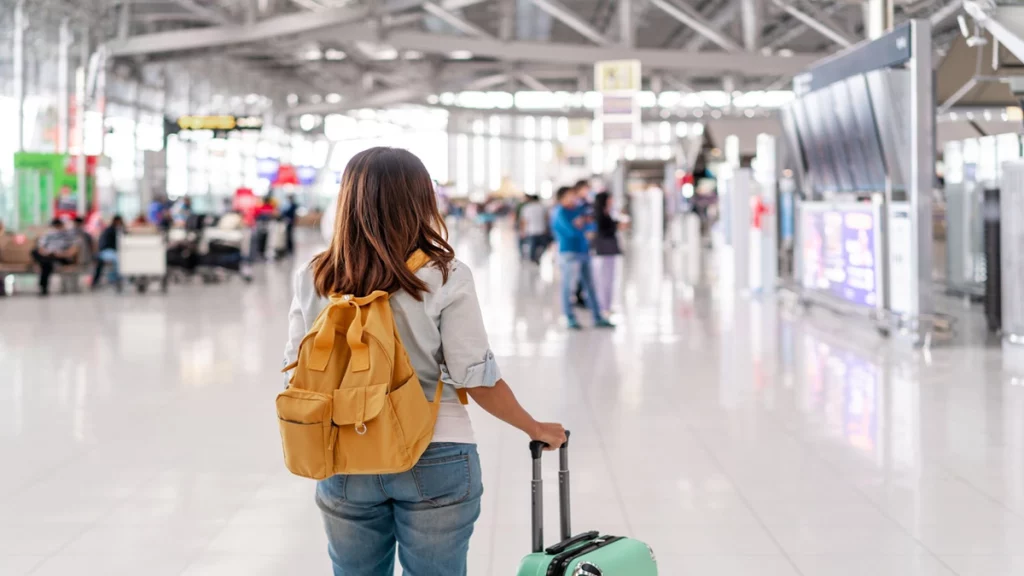 Mulher de costas com mochila e mala de mão ao lado. Ao fundo, um aeroporto desfocado. 