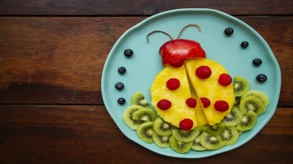 Imagem de um prato colorido infantil contendo diversos tipos de frutas, que formam o desenho de uma joaninha.