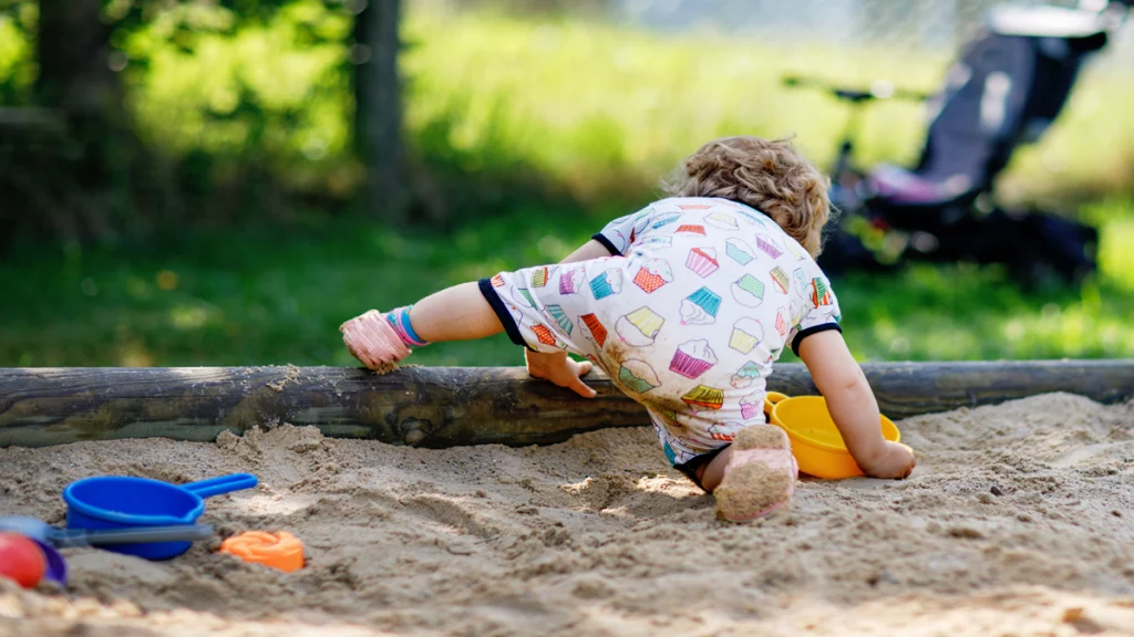 Imagem de uma criança de costas, agachada, brincando em um tanque de areia em um parque.