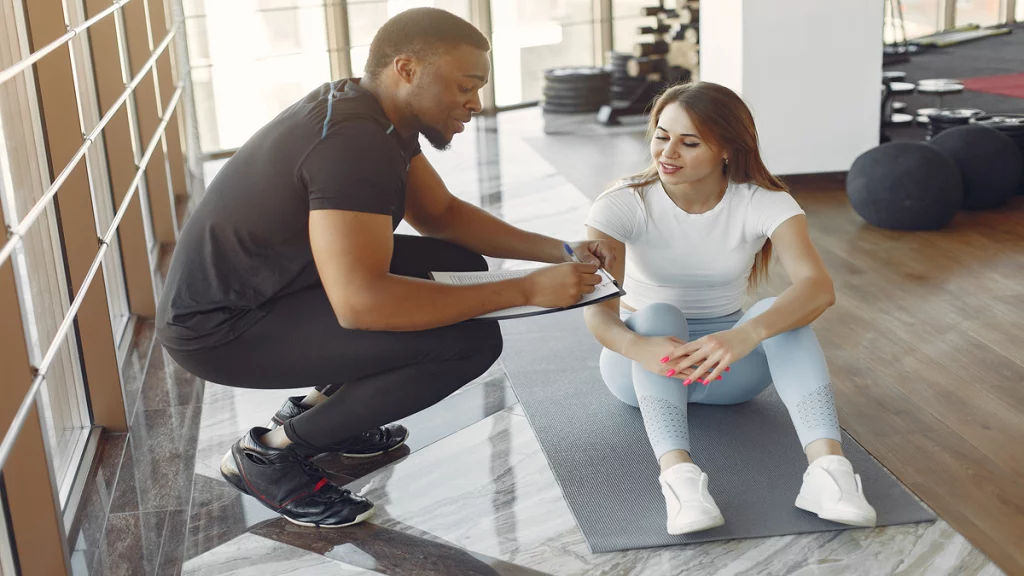 Imagem de um profissional de academia anotando informações de uma mulher durante a prática de um exercício.