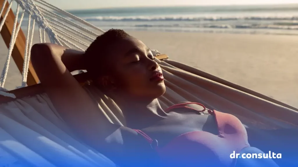 Imagem de uma mulher negra tomando sol. Ela está deitada em uma rede e em uma praia.