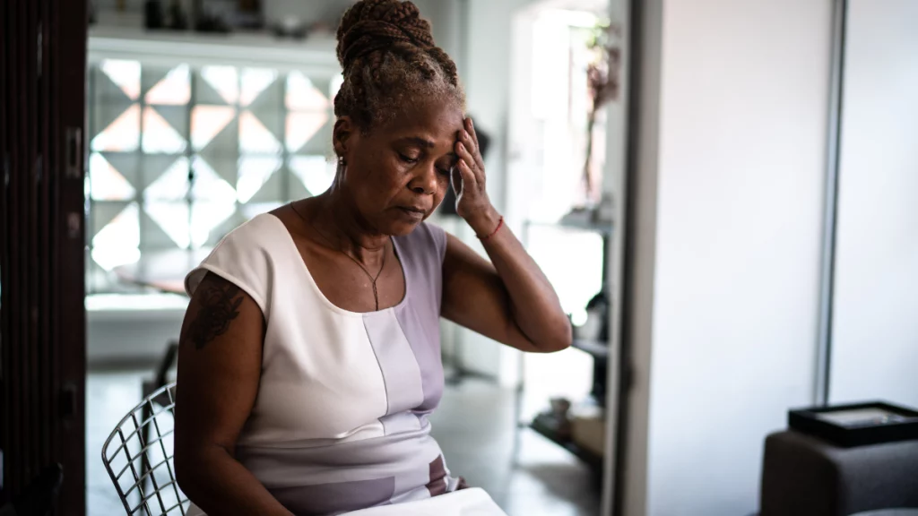 dr.consulta - mulher negra com cabelo trançado, sentada na cozinha de casa, com as mãos na cabeça, mal-estar causado pela pneumonia