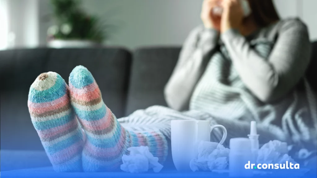 dr.consulta - mulher sentada no sofá, com roupa de frio e meia nos pés que estão sob a mesa onde é possível ver lenços de papel e uma xícara com chá, cuidados com a saúde, pneumonia