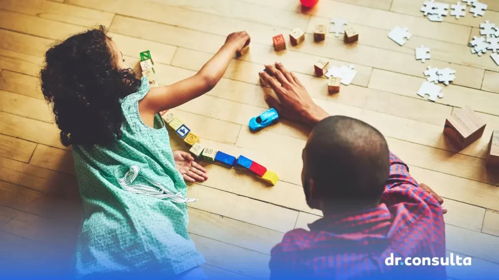 Imagem de um homem brincando com blocos e quebra-cabeça com uma menina pequena. Ambos estão deitados no chão.