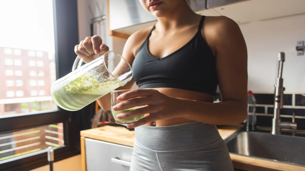 Imagem de uma mulher vestindo roupas fitness e servindo um copo de suco verde.