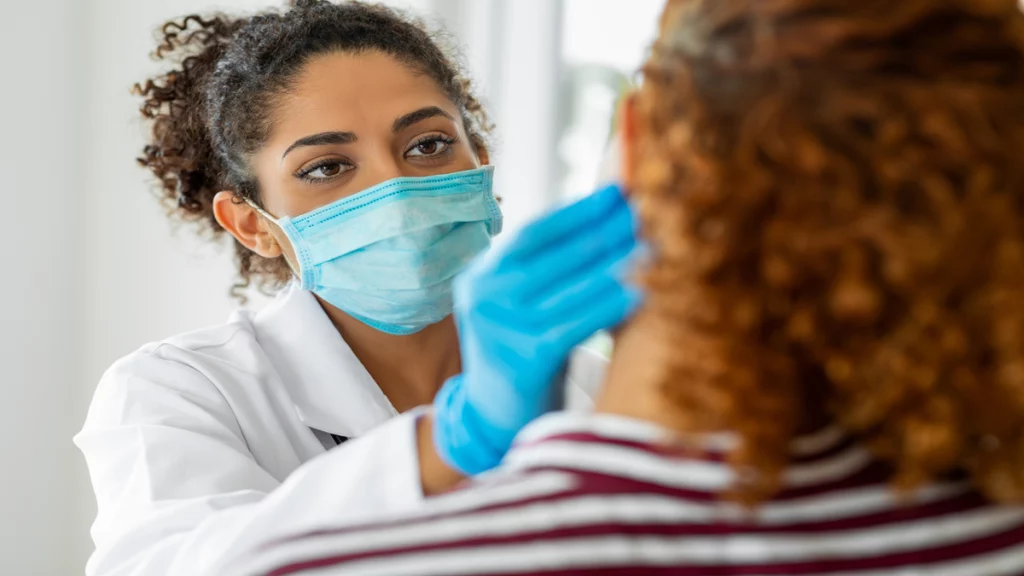 Imagem de uma médica de pele negra e cabelos castanhos escuros examinando sua paciente que está de costas para a câmera. A médica utiliza máscara, luvas cirúrgicas e um jaleco branco.