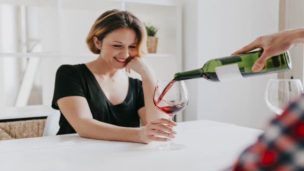 Imagem de uma mulher sorrindo sentada em uma mesa. Ela segura uma taça enquanto seu vinho é servido.