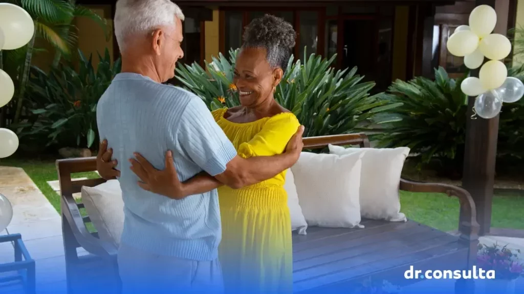 Imagem de um casal da terceira idade se abraçando e sorrindo. Ela está de vestido amarelo e ele de camisa azul.