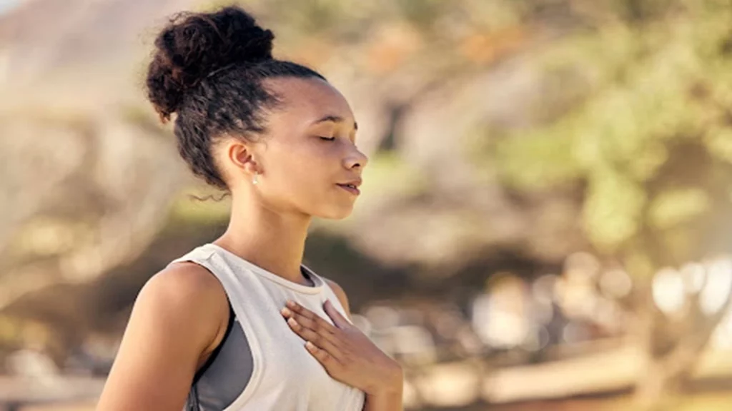 Imagem de uma mulher negra, de cabelos presos e mão sobre o peito, utilizando técnicas de respiração ao ar livre.