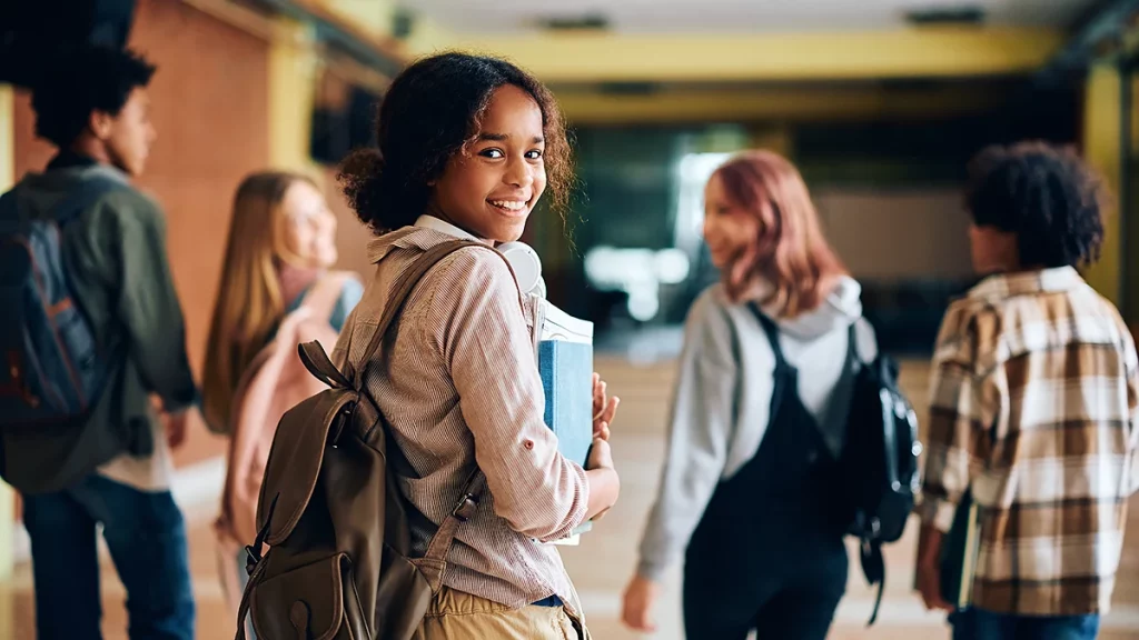 garota na fase da puberdade, carregando uma mochila nas costas e livros e apostilas nos braços, junto a um grupo de jovens que estão desfocados na imagem