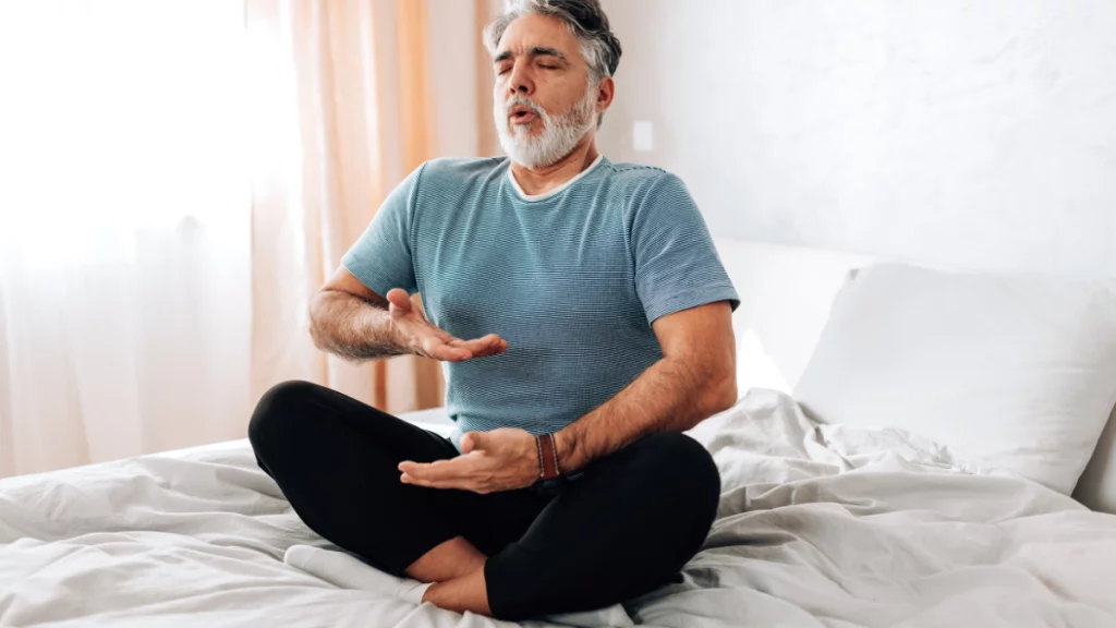 dr.consulta - homem branco de meia idade sentado na cama, fazendo um dos exercícios de yoga, prática de yoga, benefícios do yoga