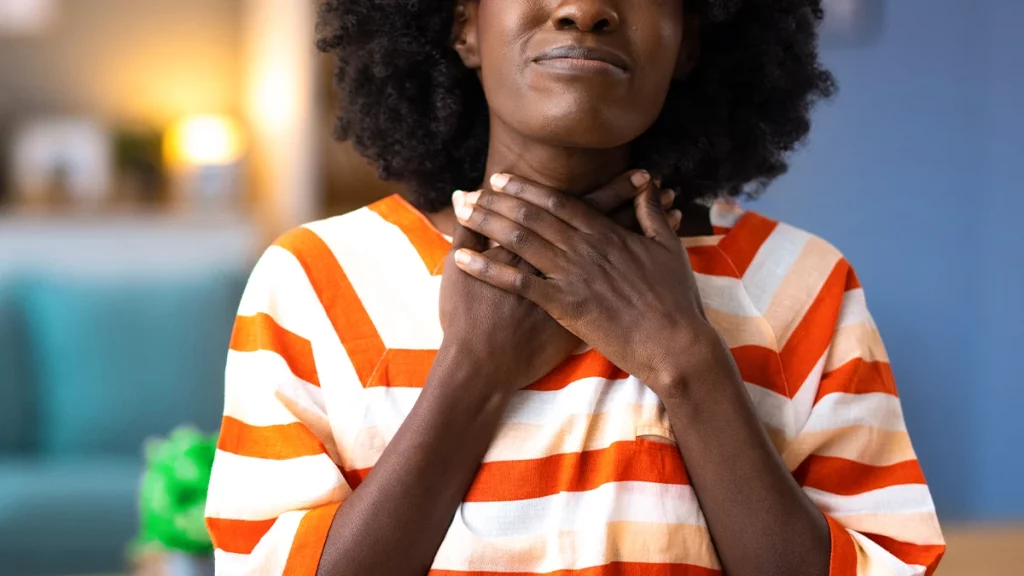 Mulher negra, com camiseta listrada laranja e branca, com as duas mãos na garganta, e com rosto indicando dor por garganta estar inflamada