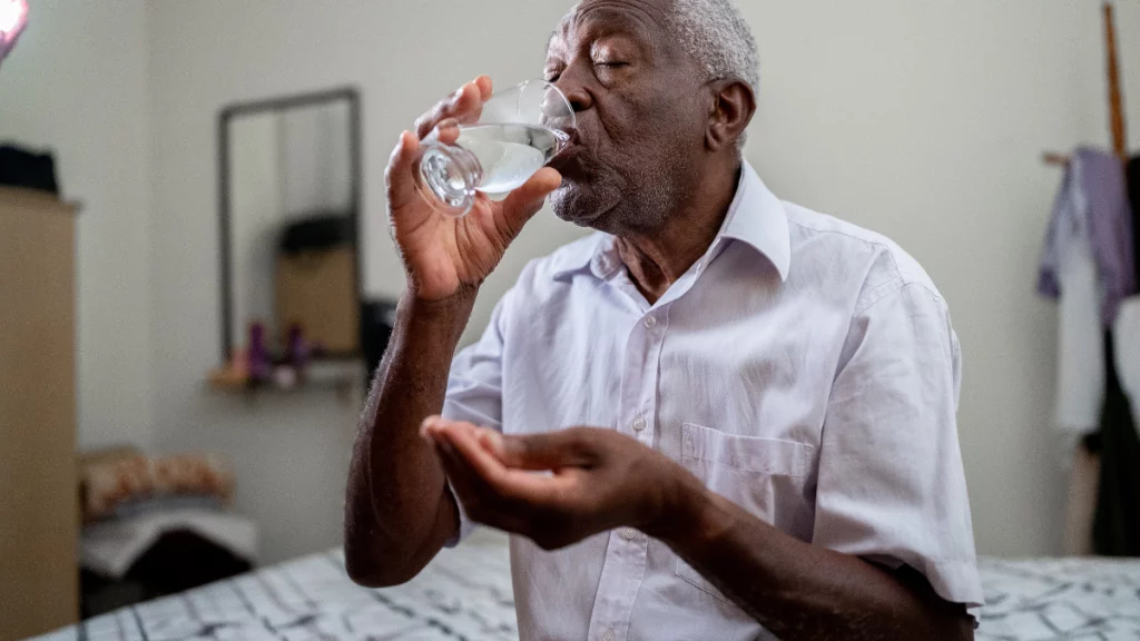 dr.consulta - homem negro da terceira sentado na beira da cama com um copo de água e um comprimido nas mãos, tomando suplemento vitamínico, vitaminas para idosos, vitaminas essenciais no cardápio, cardápio balanceado, alimentação saudável na terceira idade