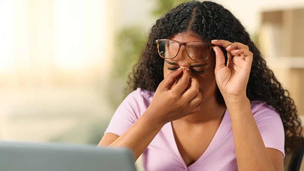 dr.consulta - mulher, em frente ao computador, segura os óculos e com a mão demonstra sentir dor atrás dos olhos