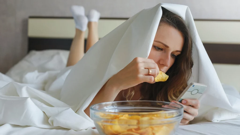 Menina comendo batatas fritas na cama, ilustrando o risco de comer antes de dormir.