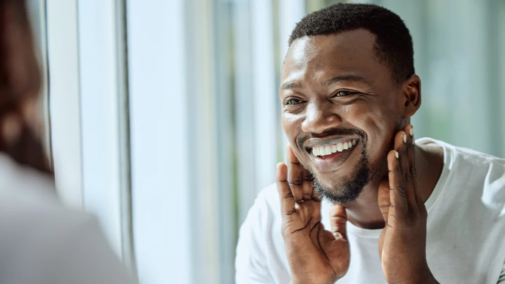 dr.consulta - homem negro se olhando no espelho com as mãos no rosto, sorrindo depois de fazer o peeling, cuidados com a pele, pele saudável e bonita, procedimento seguro