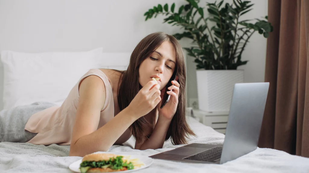 dr.consulta - pessoa na cama falando no celular e comendo hambúrguer; má alimentação e baixa exposição solar são algumas das causas para vitamina D baixa