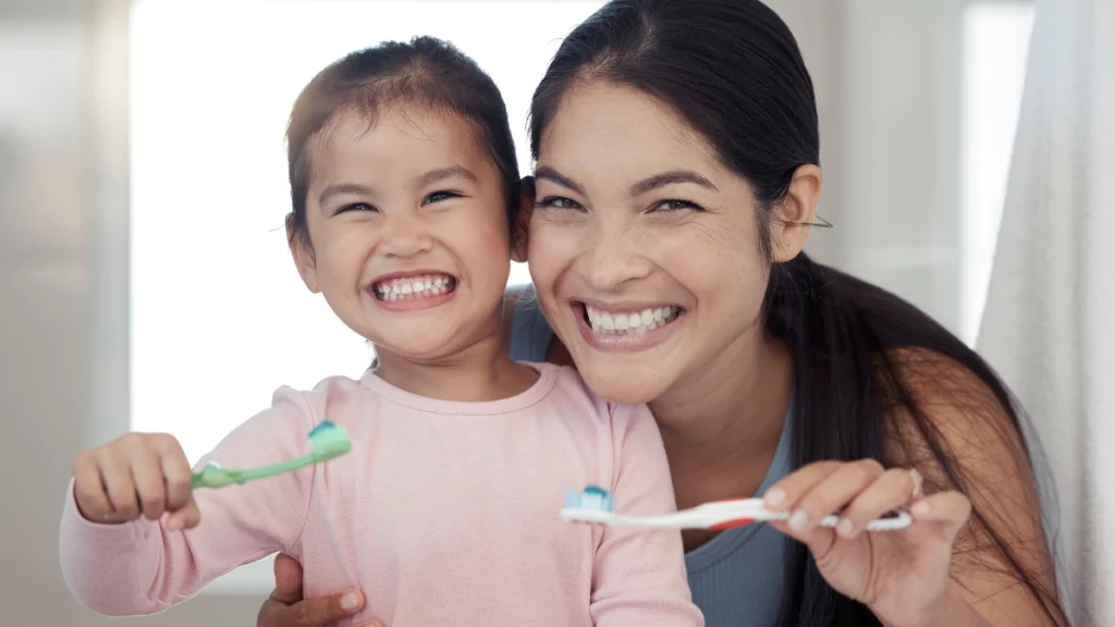 dr.consulta - Mãe e filha sorrindo. Elas seguram escovas de dente com creme dental com flúor.