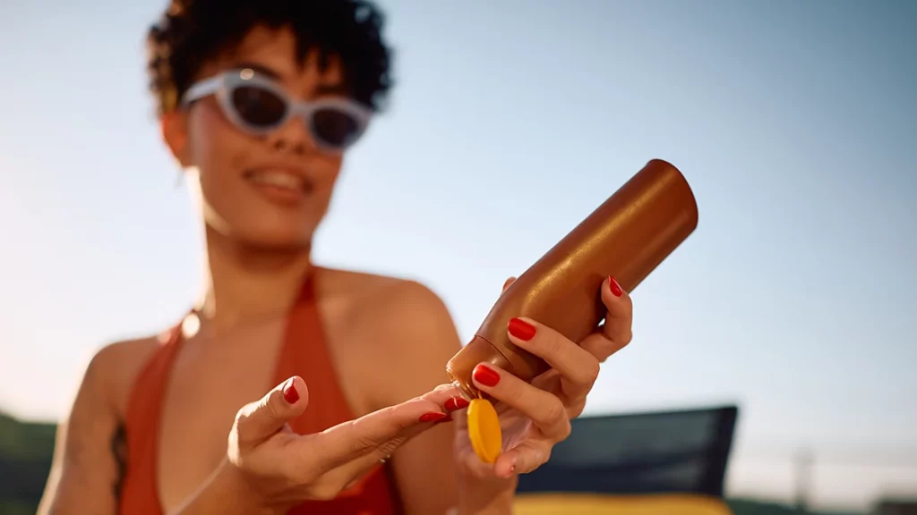 dr.consulta - Mulher sorridente ao ar livre aplicando protetor solar nas mãos para evitar queimadura de sol.