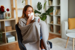 Mulher em casa enrolada em um cobertor, espirrando e segurando uma caneca quente, com sintomas de gripe ou resfriado.